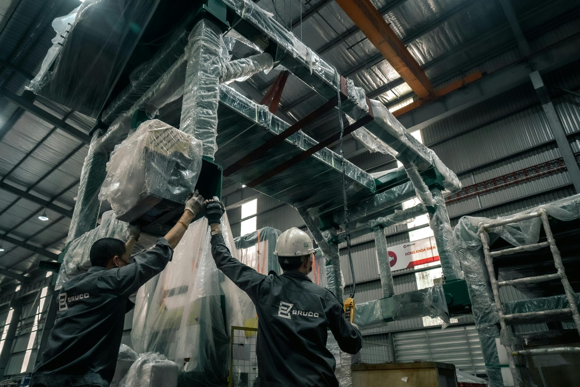 Two workers install large industrial machinery in a warehouse. They wear helmets and work with precision.
